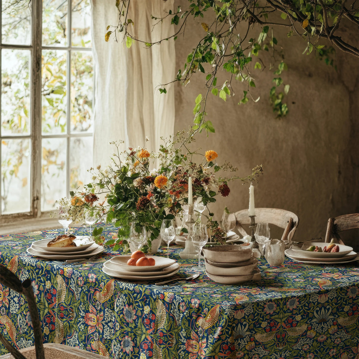Strawberry Thief tablecloth in Country Blue on a candlelit table beside a sunlit window, set with linen, stoneware and garden flowers