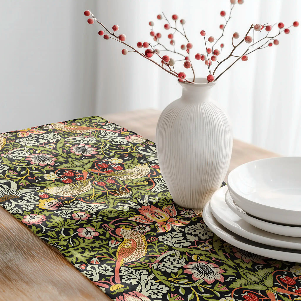 Decorative table setting with William Morris table runner featuring Strawberry Thief pattern with black base, white vase with branches, and white plates.