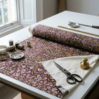 A roll of William Morris Blackthorn fabric in Maroon & Mauve part-unrolled on a work table with scissors and thread.