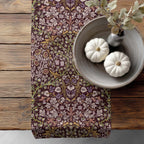 William Morris Blackthorn table runner in maroon and mauve, with floral pattern on a wooden surface with pumpkins and a vase.