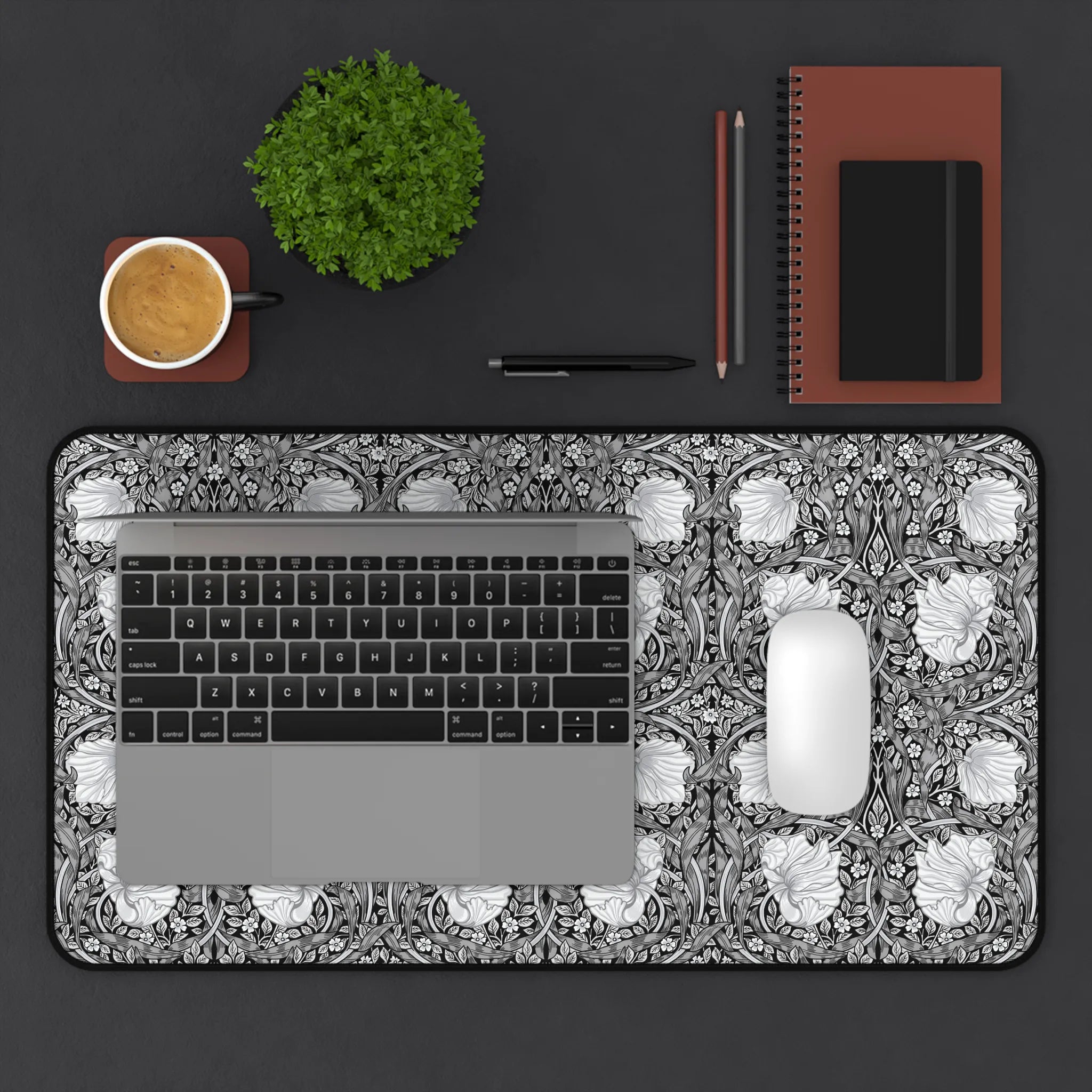Laptop on a decorative William Morris desk mat with a cup, plant, notebook, and pen on a dark surface