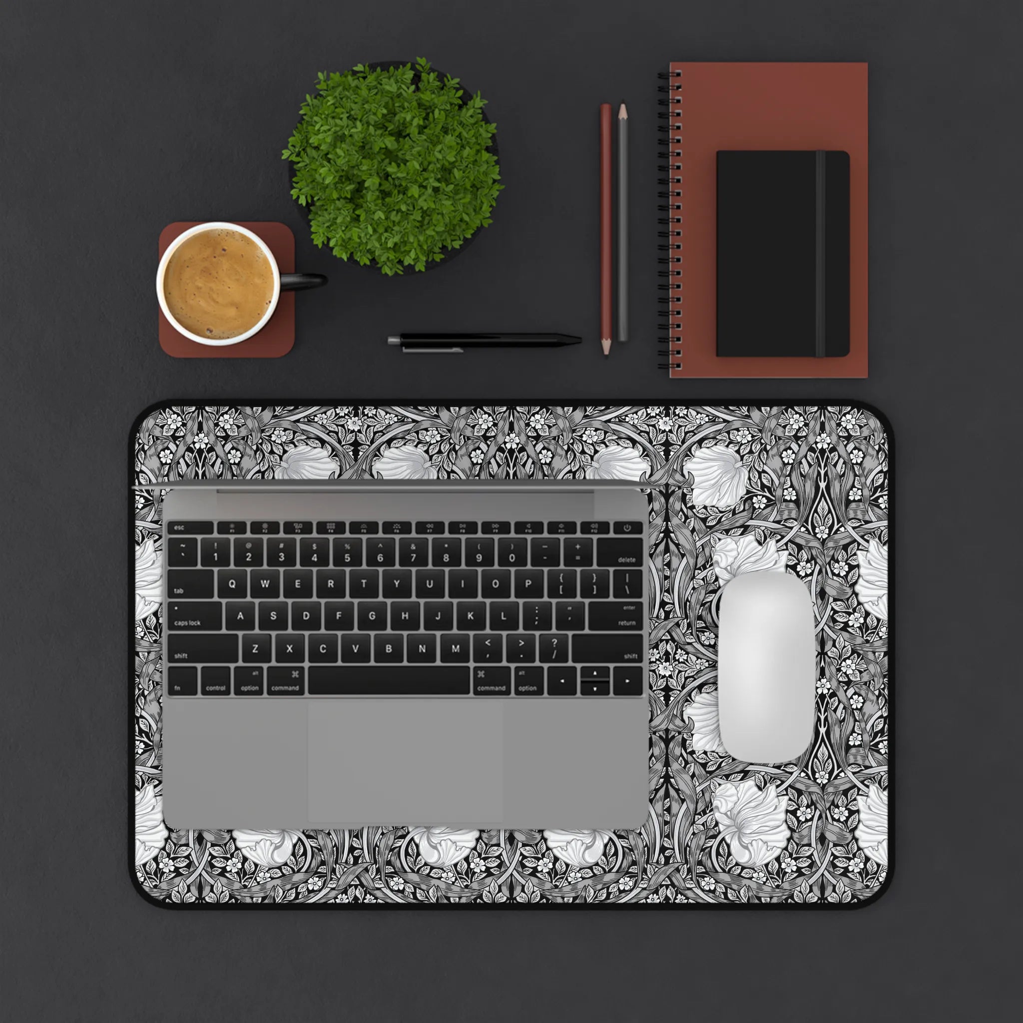 Laptop on a decorative William Morris desk mat with a cup, plant, and office supplies on a dark surface