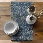 Decorative table setting with William MOrris blue table runner, floral patterned, white plates, and a vase on a wooden table.