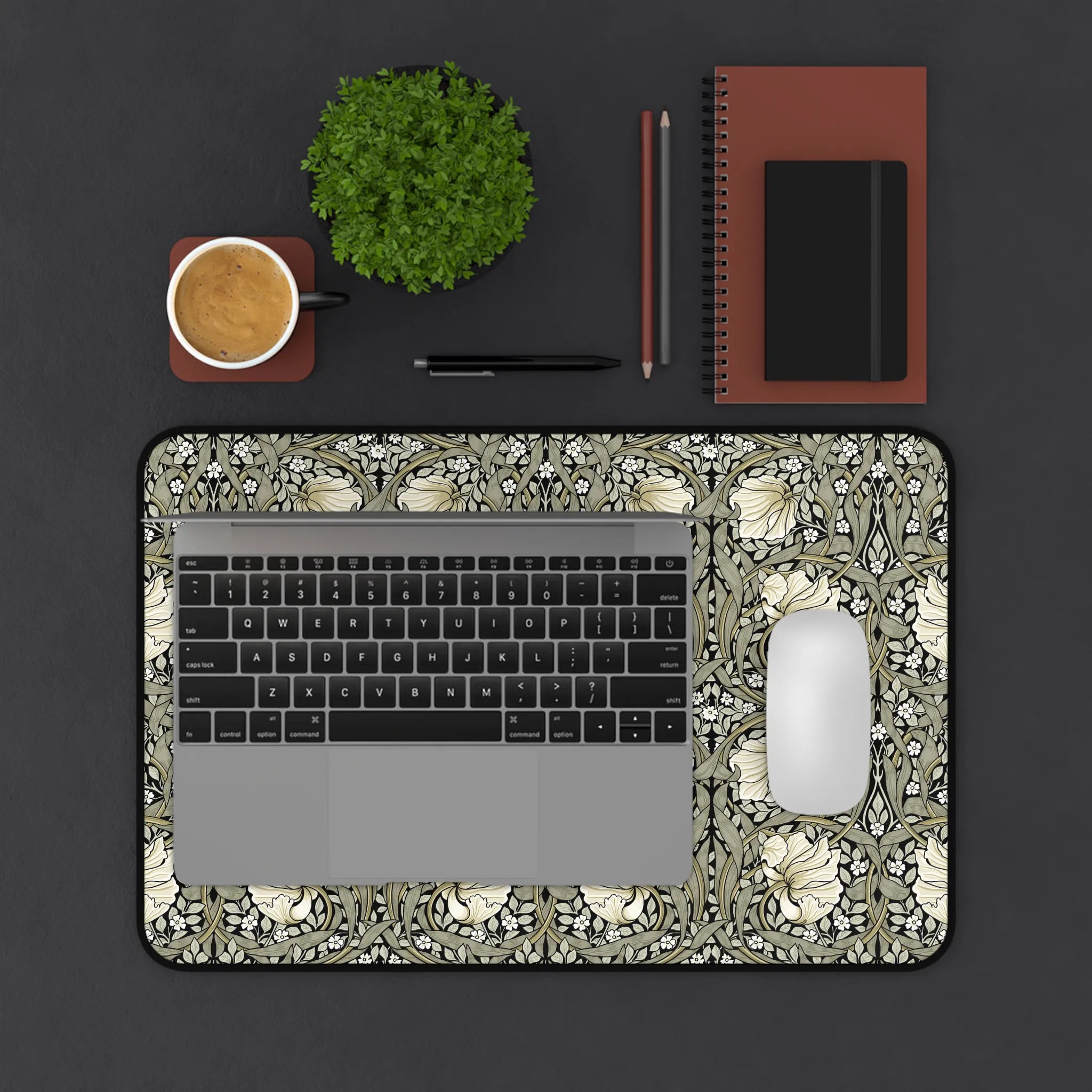Laptop on a William Morris Pimpernel desk mat in Sage and Cream Pimpernel, on top of a dark surface with a cup, plant, and stationery items.