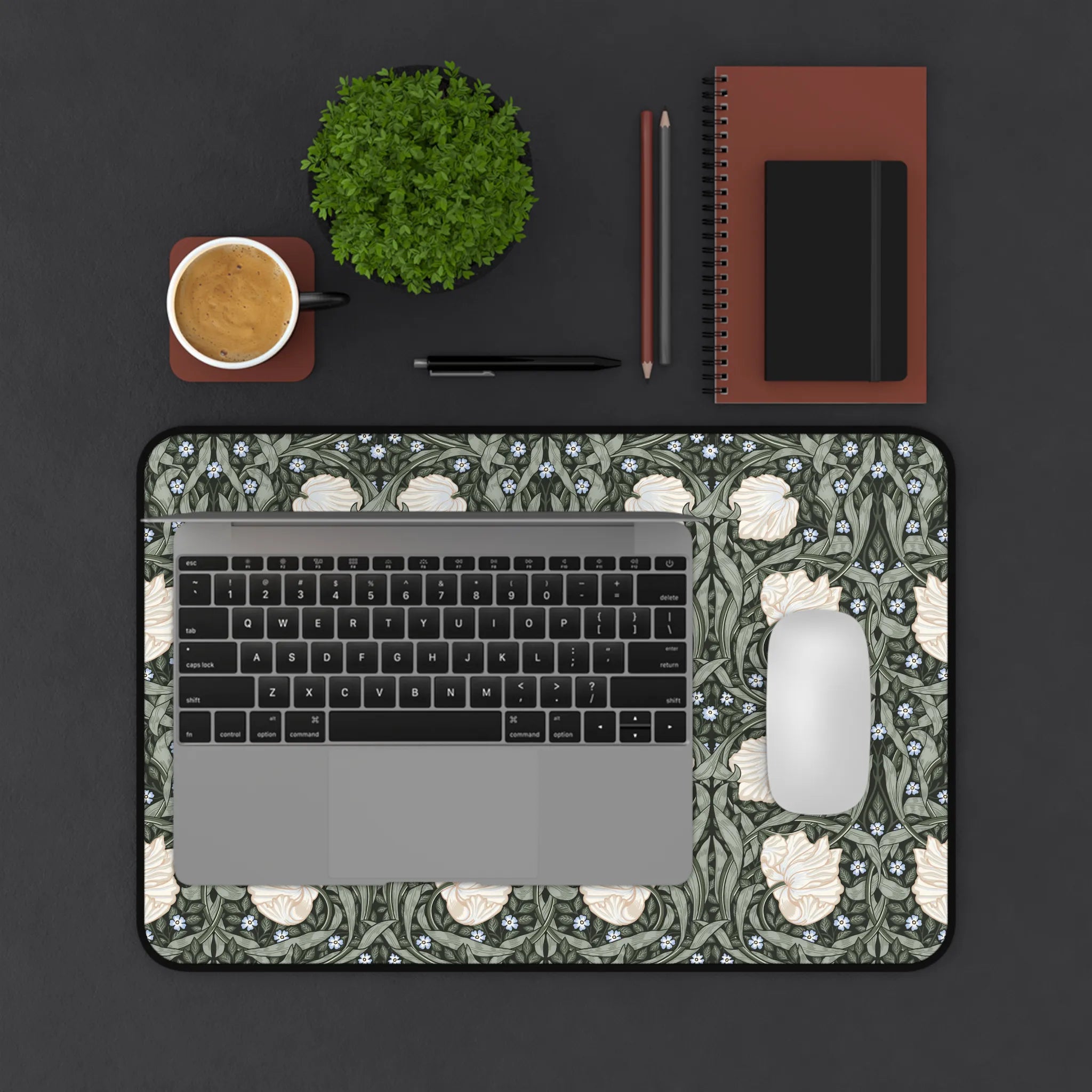 Laptop on a decorative William Morris mouse pad with a cup, plant, and stationery items on a dark surface