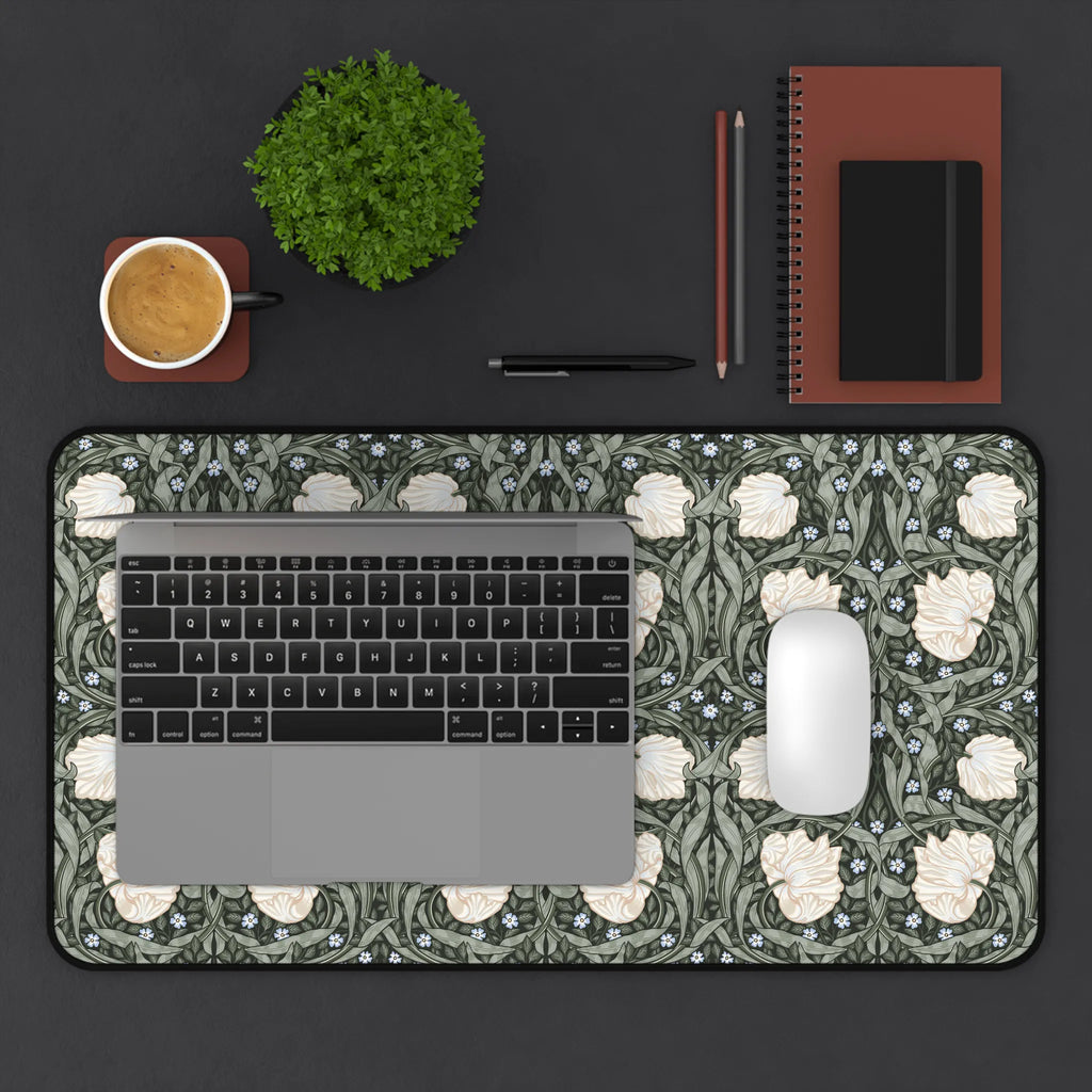 Laptop on a decorative William Morris desk mat mouse pad with a cup, plant, pen, and notebook on a dark surface