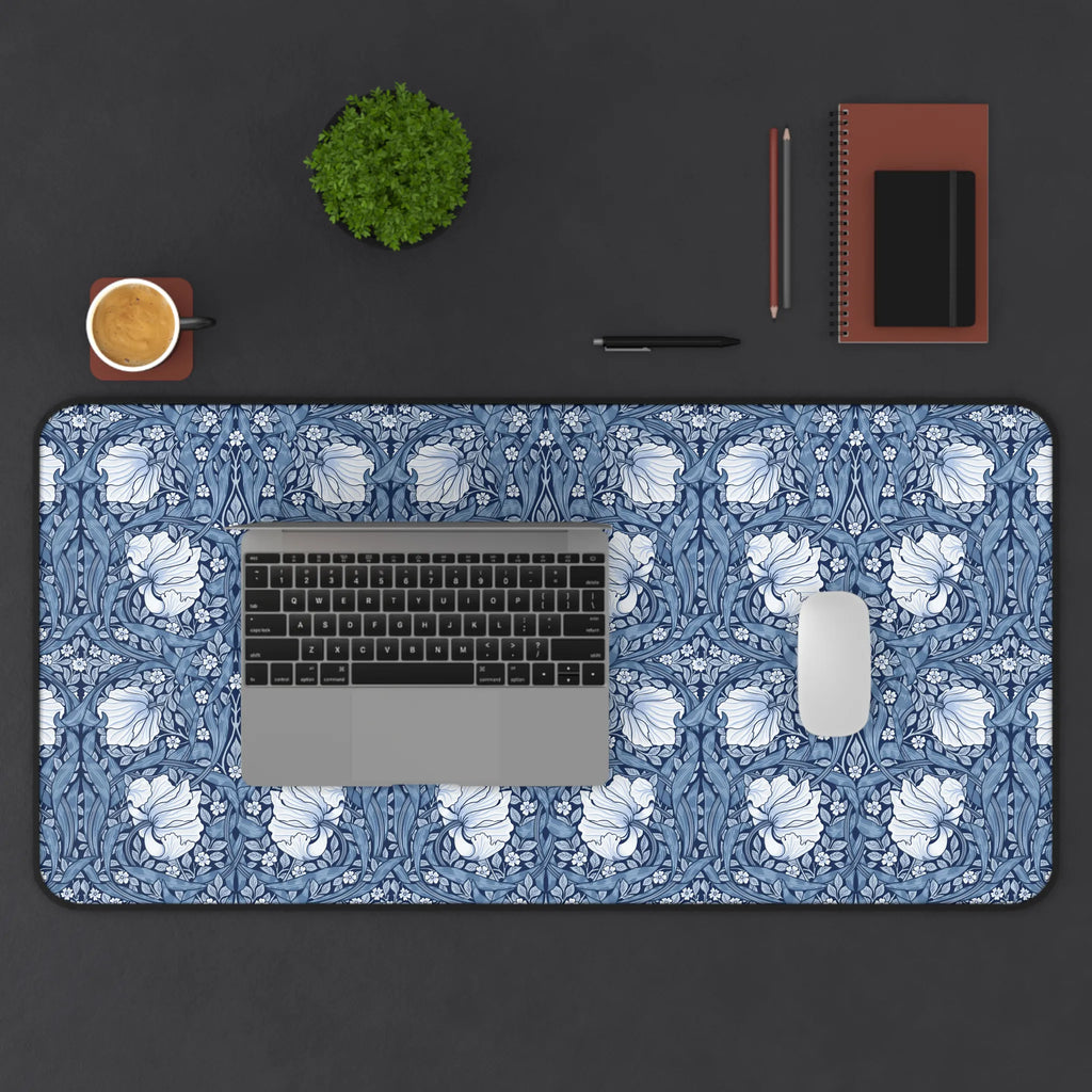 Desk setup with a blue Pimpernel by William Morris desk mat, laptop, mouse, cup, and stationery items on a dark surface.