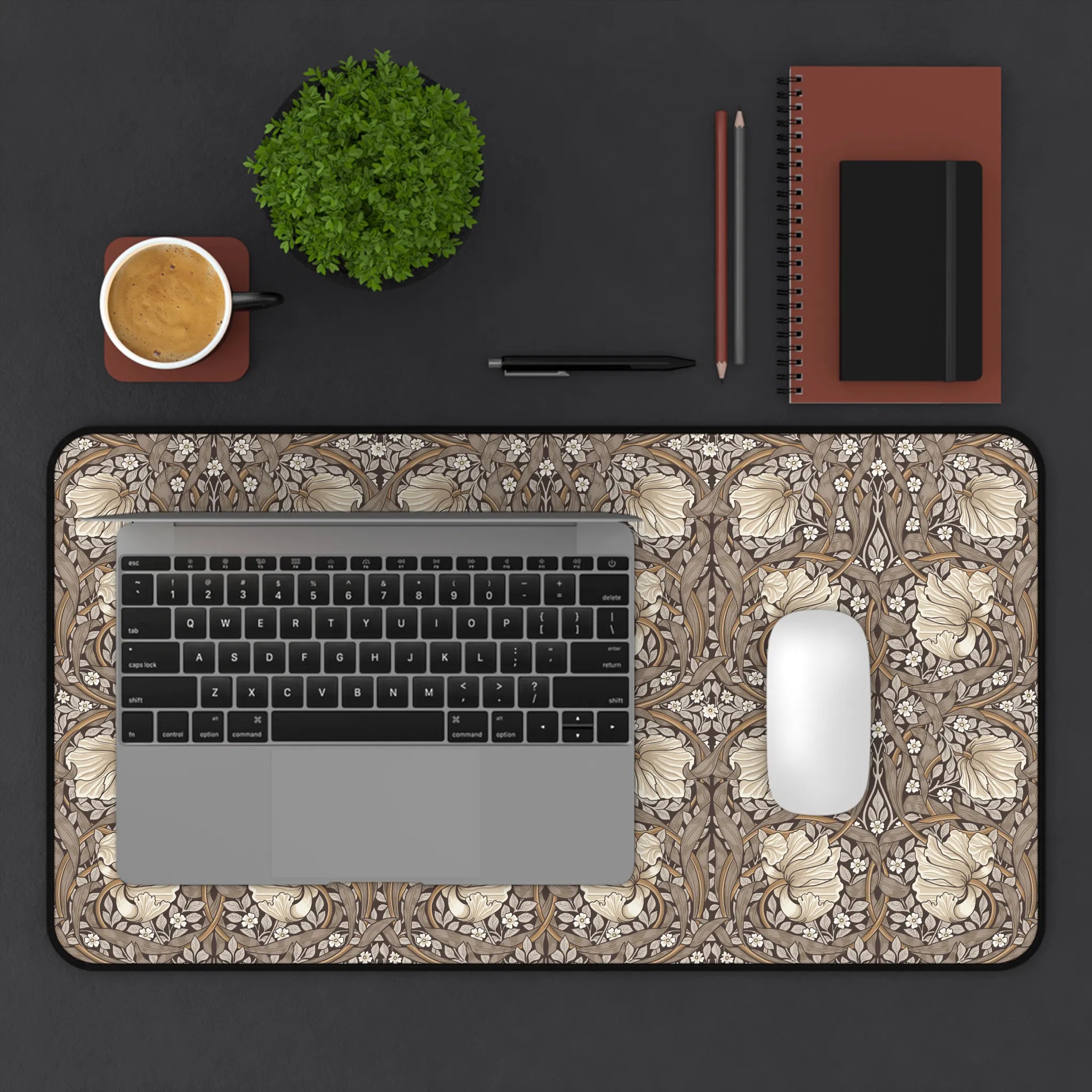 Laptop on a decorative William Morris desk mat with a cup, plant, pen, and notebook.